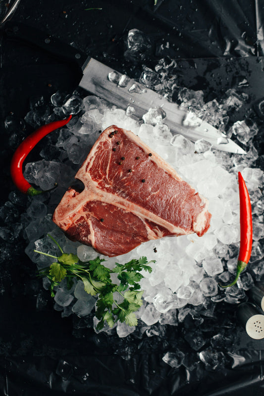 Raw steak with black peppercorns placed on a bed of ice, surrounded by red chili peppers, parsley, and a large chef’s knife on a dark surface.